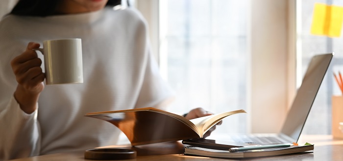Cropped image of beautiful woman holding a coffee cup in hand while reading a book and sitting in front her computer laptop that putting on wooden table over comfortable living room as background.