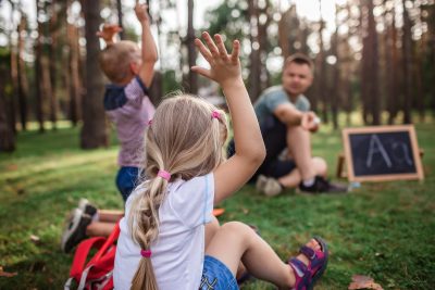 New normal back to school. Kindergarten and elementary scholars sitting with teacher on grass at open air class during pandemic. Safe hybrid education, social distance rule, new schooling guidance