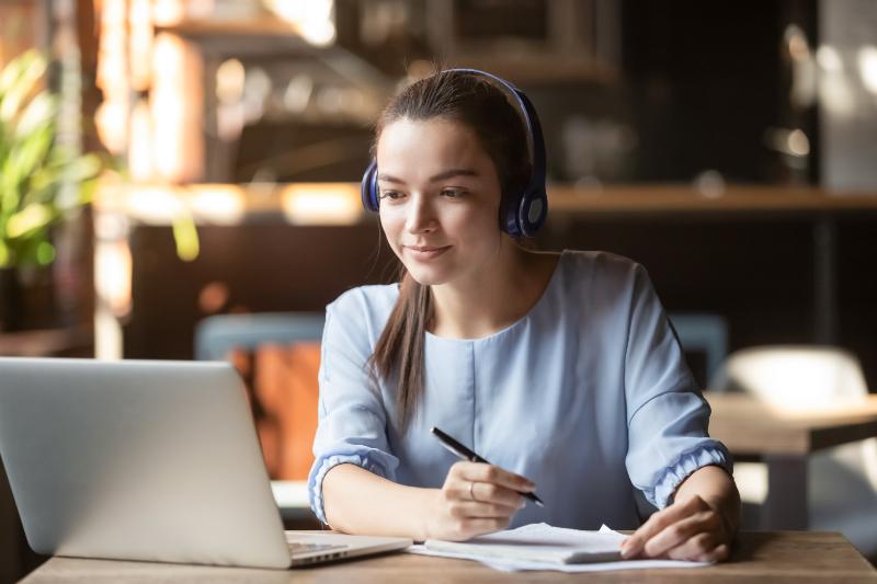 Webinar_Leading_a_Team_Remotely_Image Focused woman wearing headphones using laptop writing notes